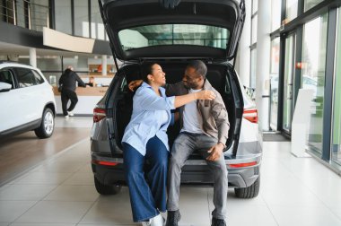 Heyecanlı çift açık bagajda oturuyor, yeni arabalarıyla hayatı kucaklıyor ve görselleştiriyor, ortak bir yolculuğu ve birlikte önemli bir alışveriş yapmanın sevincini simgeliyor.