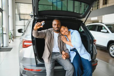 Heyecanlı çift yeni araba anahtarlarını gösterirken araba galerisindeki açık sandıkta oturup alışverişlerini kutluyorlar.