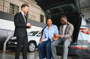 Mutlu çift araba bagajında otururken, satış elemanı modern galeride yeni araç özelliklerini gösteriyor.