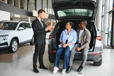 Profesyonel araba satıcısı, açık bagajda oturan Afro-Amerikan çifte yeni bir araç sunuyor. Modern galeride alım opsiyonlarını tartışıyorlar.