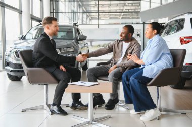 Araba satıcısı bir çiftle tokalaşıyor ve modern bir galeride yeni bir araba alıyor.