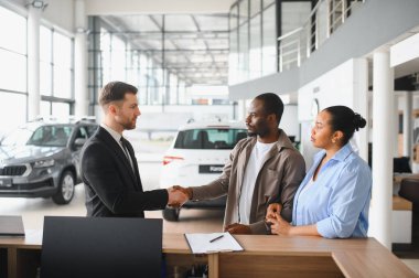 Ortağı bakarken araba satıcısı bir müşteriyle tokalaşıyor ve modern bir araba galerisinde başarılı bir anlaşmayı kutluyor.