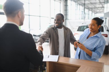 Heyecanlı çift bir satıcıyla el sıkışıyor, galeride yeni arabalarını satın alıyorlar, başarılı bir anlaşmayı kutluyorlar.