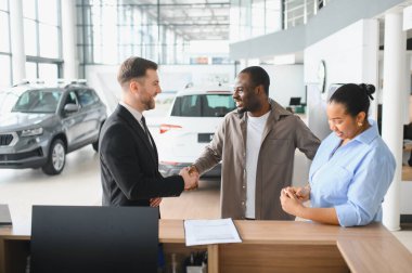 Mutlu satıcı bir çiftle el sıkışıyor, galeride yeni bir araba almalarını kutluyor.