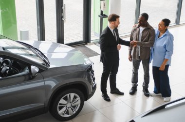 Satıcı neşeli bir çiftle el sıkışıyor. Parlak ve modern bir galeride yeni araba alımlarını kutluyorlar.