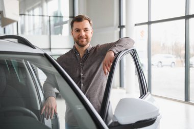 Gülümseyen adam galeride yeni bir araba seçiyor, galeride açık bir araca yaslanıyor.