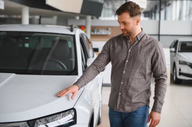 Bir galeri içinde yeni bir beyaz arabanın kaputuna dokunan bir adam.