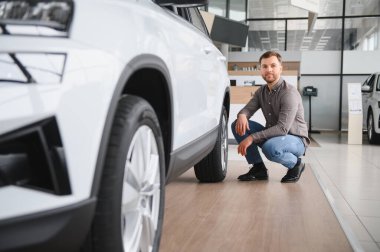 Bir araba galerisinde beyaz bir cipin yanında çömelmiş duran bir adam satın almadan önce aracı inceliyor.