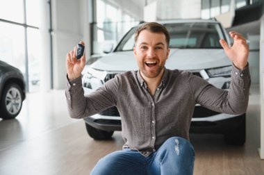 Hevesli bir adam araba anahtarlarını tutuyor, modern bir araba galerisinde aldığı yeni arabayı kutluyor, neşe ve memnuniyet sergiliyor.