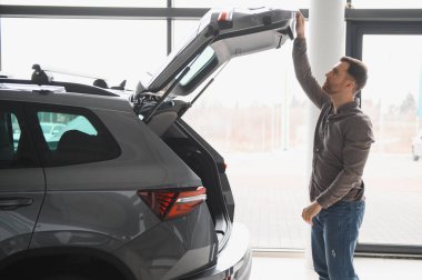 Bir araba galerisini ziyaret ederken yeni bir aracın bagaj alanını inceleyen adam, satın alma seçeneklerini değerlendiriyor