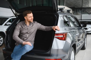 Gülümseyen adam rahat bir şekilde yeni bir cipin bagajında oturuyor, geniş özelliklerini ve tasarımlarını modern bir araba galerisinde inceliyor.