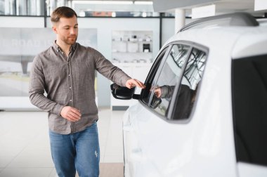 Müşteri galeride yeni bir beyaz arabayı inceliyor, pencereye dokunuyor ve alışverişini düşünüyor.