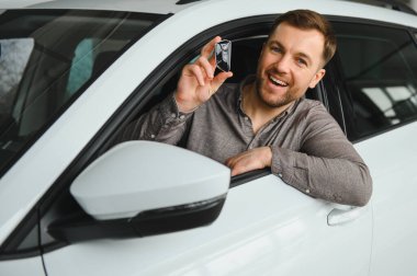 Hevesli bir adam arabada oturuyor, elinde gülümsemeli bir anahtar tutuyor, modern bir galeride yeni arabası için heyecanını sergiliyor.