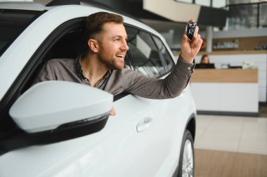 Gülümseyen müşteri yeni bir araçta otururken arabanın anahtarlarını tutuyor galerinin içinde, neşe ve heyecan saçıyor.