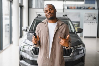 Gururlu Afro-Amerikalı adam araba anahtarlarını gösterirken bir araba galerisini işaret ederek satın alışını kutluyor.