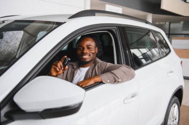 Afrika kökenli Amerikalı müşteri araba anahtarlarını tutuyor, yeni arabasında oturuyor, gülümsüyor, galerinin içinde.