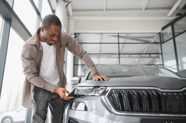 Adam araba galerisinde parlak gri bir arabanın farını inceliyor. Heyecan ve gururla potansiyel bir alışveriş düşünüyor.