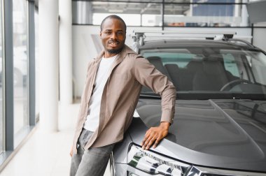 Gülümseyen adam araba galerisinde yeni arabasına yaslanıyor, son alışverişinin tadını çıkarıyor.