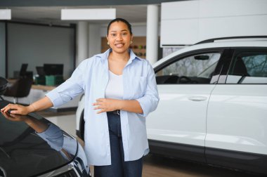 Afro-Amerikalı bir kadın, canlı bir araba galerisinde yeni bir araba seçiyor, parlak bir arabanın kaputuna güvenle dokunuyor.