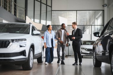 Satıcı Afrikalı Amerikalı çifte yeni arabaları gösteriyor. Bir araba galerisinde doğru aracı seçmelerine yardım ediyor.