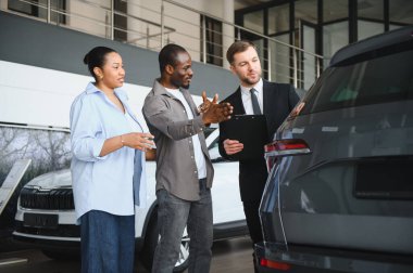 Satış elemanı Afro-Amerikan çifte yeni araba özelliklerini gösteriyor. Araba galerisinde alışverişi tartışıyorlar.