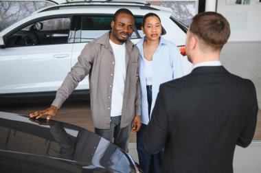 Afro-Amerikan çift yeni bir araba seçiyor, araçları inceliyor ve bir satıcıya danışıyor.