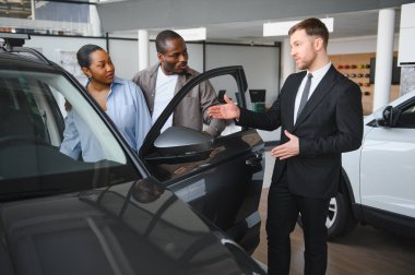 Araba satıcısı, modern bir galeride Afro-Amerikan bir çifte yeni bir arabanın özelliklerini sunuyor.