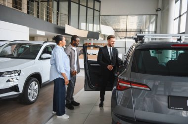 Modern bir galeride Afro-Amerikan bir çifte yeni bir arabanın özelliklerini gösteren profesyonel bir araba satıcısı.