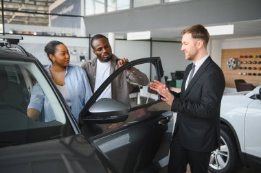 Araba satıcısı, yeni bir arabanın özelliklerini galeriden bir araç almak isteyen Afrikalı Amerikalı bir çifte sunuyor.