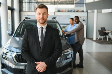 Profesyonel araba satıcısı modern bir galeride güven içinde poz verirken bir çift heyecanla yeni araçlarını tartışıyor ve seçiyor.