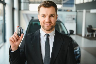 Gülümseyen araba satıcısı araba anahtarlarını bir galeride tutuyor, modern bir galeride yeni bir araç almanın heyecanını tanıtıyor.