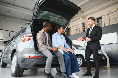 Satıcı, bir galeride yeni bir arabanın bagajında oturan bir çifte araba sunuyor.