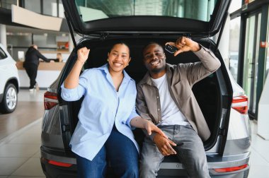 Afro-Amerikan çift yeni arabalarında oturuyor, gülümsüyor ve anahtarlarını tutuyorlar, arka planda bir satıcı var.