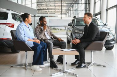 Bir çiftin önünde oturan araba satıcısı, modern bir galeride yeni bir araba teklifini tartışıyor.