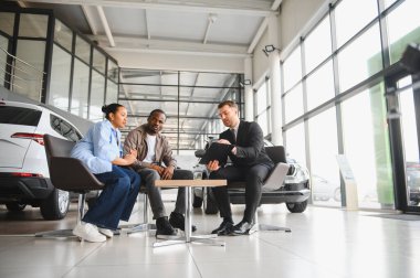 Araba satıcısı, bir araba galerisindeki masada oturan Afrikalı Amerikalı çifte satış sözleşmesini açıklıyor.