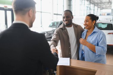 Gülümseyen Afro-Amerikan çifti araba satıcısıyla el sıkışıyor, modern sergi salonunda alışverişi bitiriyor.