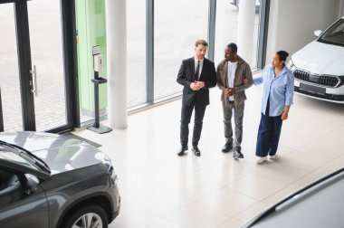 Araba satıcısı, modern bir araba galerisini ziyaret eden Afro-Amerikan bir çifte yeni araçlar sunuyor.