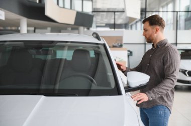 Adam galeride yeni beyaz arabayı kontrol ediyor, arabaya dokunuyor ve yeni aracını seçiyor.