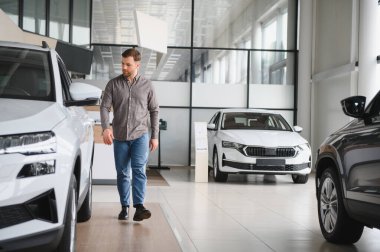 Modern bir araba galerisinde yürüyen adam, çeşitli model ve seçenekleri incelerken düşünceli bir şekilde yeni bir araç seçiyor.