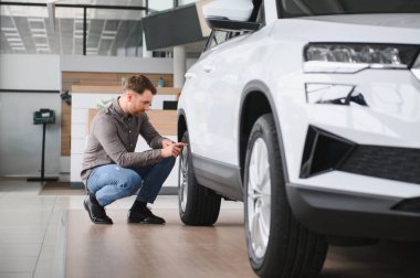 Yeni bir araba galerisinde çömelmiş yeni bir cipin lastiklerini inceleyen bir adam, satın alma seçeneklerini göz önünde bulundurarak...