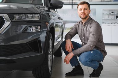 Gösterişli, modern bir arabanın yanında diz çökmüş genç bir adam, onun özelliklerini ve tasarımlarını değerlendiriyor, karar verme sürecini yansıtıyor.