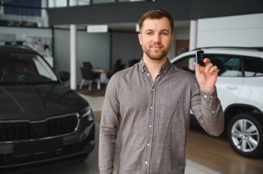 Yeni bir araç satın almayı bitirdikten sonra galeri galerisinde araba anahtarını gururla sergileyen mutlu müşteri neşe ve memnuniyet saçıyor.