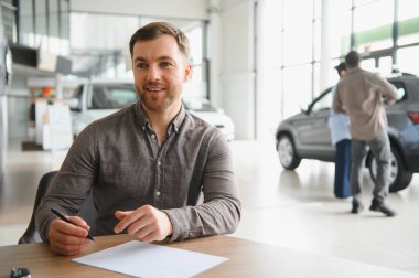 Araba satıcısı elinde bir kalem tutuyor ve bir araba galerisinde masasında otururken bir çift yeni bir araç seçiyor.