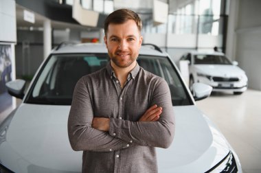 Yeni bir arabanın önünde kollarını kavuşturmuş, modern bir galeride satış yapmaya hazır gülümseyen bir müşterinin portresi.