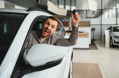 Yeni araba alımını kutlayan heyecanlı bir adam, lüks araçlarla dolu modern bir galeride otururken gururla anahtarlarını sergiliyor.