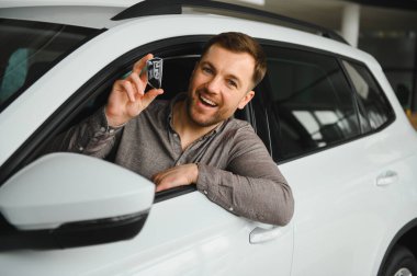 Araba anahtarlarını gösteren heyecanlı bir adam yepyeni beyaz bir arabanın içinde oturmuş bir araba galerisinde alışverişini kutluyor.