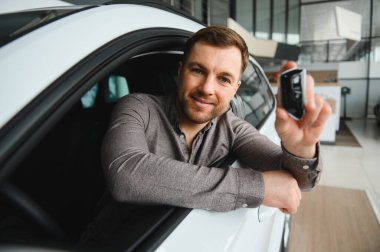 Adam lüks bir galeride arabayı inceliyor, elinde anahtarlar var ve satın almayı düşünüyor. Ayarlar modern ve profesyonel bir çevreyi yansıtıyor.