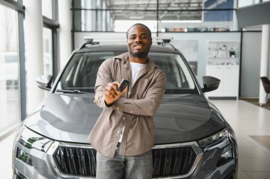 Gülümseyen müşteri gururla araba anahtarını galerideki yeni aracın önünde sergiliyor, başarılı alım ve mülkiyeti kutluyor.