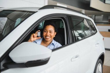 Gülümseyen Afrikalı Amerikalı kadın yeni beyaz bir arabanın içinde otururken araba anahtarlarını gösteriyor. Modern bir bayiden aldığı için mutlu.