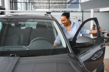 Bir galeride yeni bir arabanın içini incelerken gülümseyen mutlu bir Afro-Amerikan kadını satın alma deneyiminin tadını çıkarıyor.